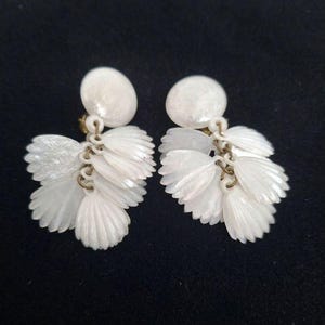 Könnte beinhalten: Ein Paar Ohrringe aus weißen Muscheln mit einem runden Stecker und mehreren hängenden, fächerförmigen Muschelsegmenten, die durch kleine messingfarbene Metallringe verbunden sind. Die Muscheln haben eine perlmuttartige, schimmernde Oberfläche.