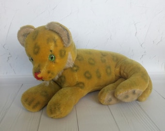 Könnte beinhalten: Ein gelber Plüschleopard mit grünen Augen und einer rosa Nase. Der Leopard liegt auf dem Bauch mit ausgestreckten Pfoten.