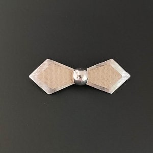May include: A silver bow tie brooch with a textured design and a round, silver ball in the center.