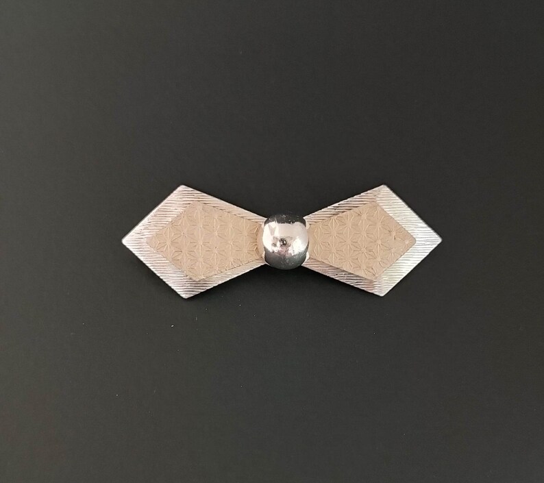 May include: A silver bow tie brooch with a textured design and a round, silver ball in the center.