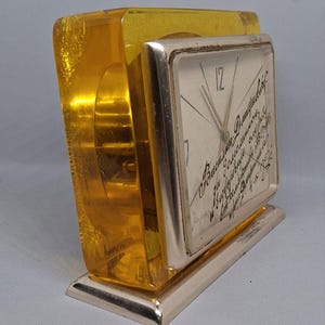 May include: A vintage desk clock with a rectangular face and a gold-colored metal base. The clock face has a light background with black hands and text. The clock is encased in a transparent, amber-colored acrylic block.