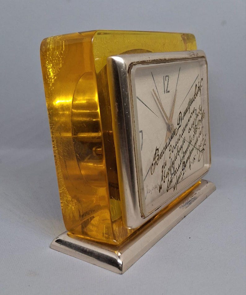 May include: A vintage desk clock with a rectangular face and a gold-colored metal base. The clock face has a light background with black hands and text. The clock is encased in a transparent, amber-colored acrylic block.