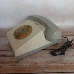 May include: A vintage rotary dial phone with a gray and white color scheme. The phone has a black cord and is sitting on a wooden surface.