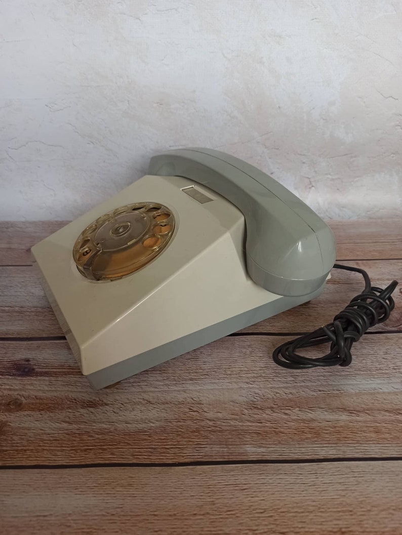 May include: A vintage rotary dial phone with a gray and white color scheme. The phone has a black cord and is sitting on a wooden surface.