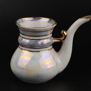 May include: A white ceramic pipe-shaped decorative object with a pearlescent finish. It features gold accents on the rim, handle, and spout. The body has a bulbous shape with a cylindrical neck and a curved spout.