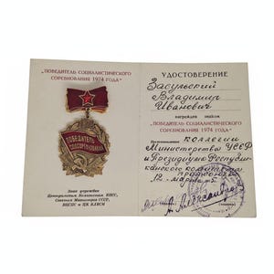 May include: A vintage award certificate and medal. The certificate is off-white with text in Russian. The medal is red and gold, featuring a star and the year 1974. The certificate includes a stamp and signature.