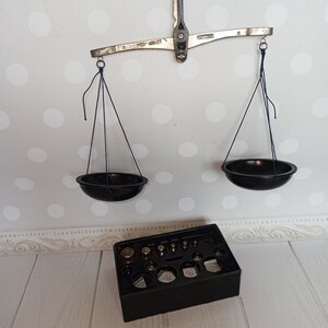 Puede incluir: Una balanza de equilibrio de plata con dos cuencos negros colgando de ella. La balanza tiene un brazo de metal con las palabras "100 GRAMS" impresas en &eacute;l. Una caja negra con un juego de pesas est&aacute; en primer plano.