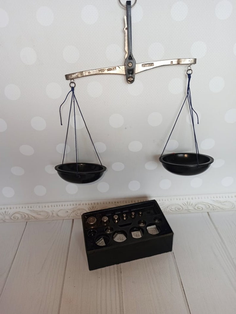 Puede incluir: Una balanza de equilibrio de plata con dos cuencos negros colgando de ella. La balanza tiene un brazo de metal con las palabras "100 GRAMS" impresas en &eacute;l. Una caja negra con un juego de pesas est&aacute; en primer plano.