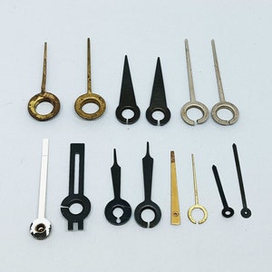Op de afbeelding: Assortiment van wijzerplaten in goud, zilver en zwart metaal. De wijzers zijn van verschillende lengtes en stijlen, waaronder recht, puntig en gebogen.