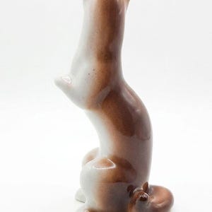 K&ouml;nnte beinhalten: Eine gl&auml;nzende Keramikfigur eines kleinen Tieres, wahrscheinlich ein Wiesel oder Frettchen, das auf den Hinterbeinen steht. Die Figur hat eine glatte, glasierte Oberfl&auml;che in Wei&szlig; und warmem Braun, einen geschwungenen K&ouml;rper und einen Ringelschwanz.