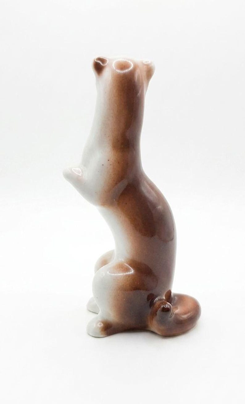 K&ouml;nnte beinhalten: Eine gl&auml;nzende Keramikfigur eines kleinen Tieres, wahrscheinlich ein Wiesel oder Frettchen, das auf den Hinterbeinen steht. Die Figur hat eine glatte, glasierte Oberfl&auml;che in Wei&szlig; und warmem Braun, einen geschwungenen K&ouml;rper und einen Ringelschwanz.