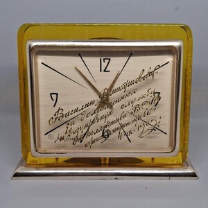 May include: A vintage rectangular clock with a silver frame and a yellow-tinted plastic casing. The clock face has black numerals and hands, with handwritten script. The clock is set on a silver base.
