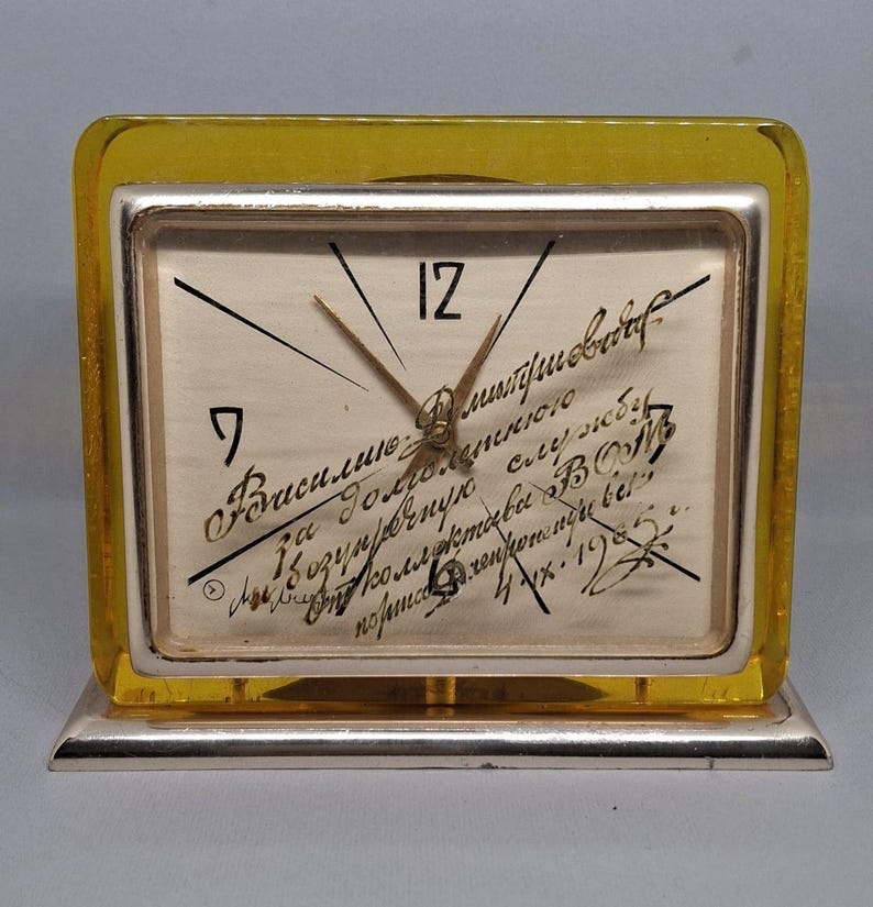 May include: A vintage rectangular clock with a silver frame and a yellow-tinted plastic casing. The clock face has black numerals and hands, with handwritten script. The clock is set on a silver base.