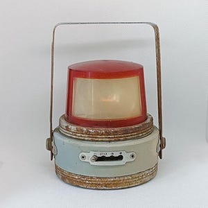 May include: A vintage, rusty, metal lantern with a red plastic shade and a handle. The lantern has a switch labeled "OTKR" and a small, round window.
