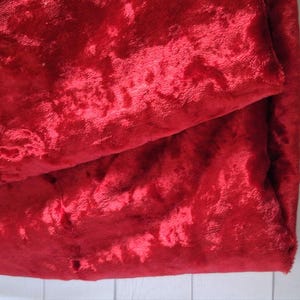 May include: A close-up of a folded piece of red velvet fabric. The fabric has a plush, textured surface with a slight sheen. The deep red color is rich and vibrant, with subtle variations in tone.