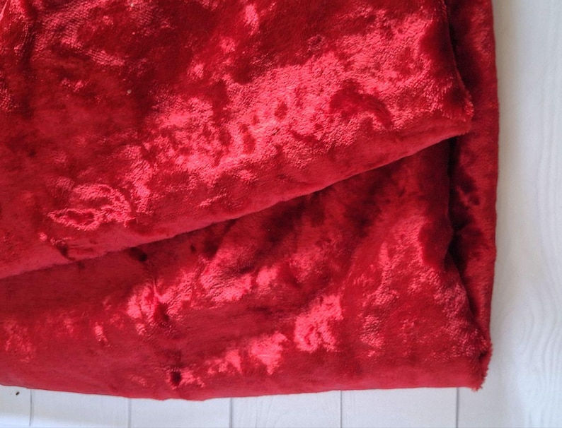 May include: A close-up of a folded piece of red velvet fabric. The fabric has a plush, textured surface with a slight sheen. The deep red color is rich and vibrant, with subtle variations in tone.