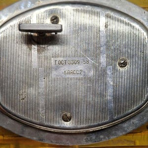 May include: Close-up of a vintage silver-toned clock mechanism. The back of the clock features a textured, vertically-lined surface with a winding key and three screws. The text "ГОСТ 8309-58 КЛАСС 2" is visible.
