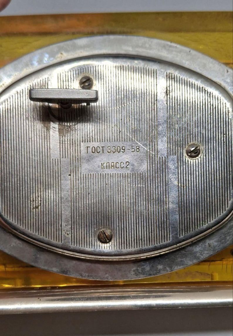 May include: Close-up of a vintage silver-toned clock mechanism. The back of the clock features a textured, vertically-lined surface with a winding key and three screws. The text "ГОСТ 8309-58 КЛАСС 2" is visible.