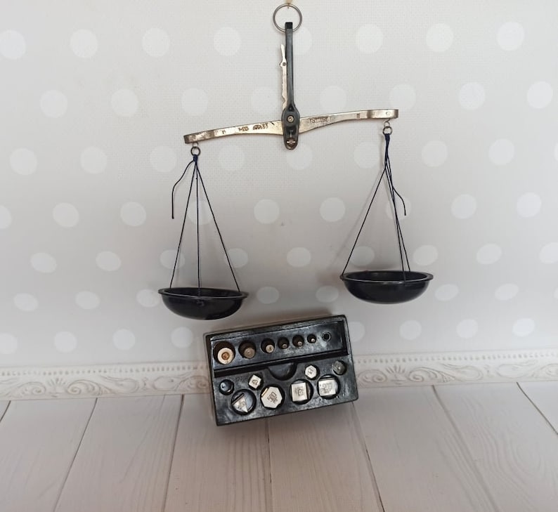 Puede incluir: Una balanza de equilibrio vintage de plata con cuencos de metal negro y un juego de pesas en una caja negra. La balanza est&aacute; colgada de una pared blanca con un patr&oacute;n de lunares.
