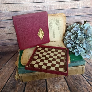 Op de afbeelding: Een vintage rode en bruine damspelset met een houten frame. Het speelbord is gesloten en de deksel ligt bovenop het bord. De spelset ligt op een stapel oude boeken.