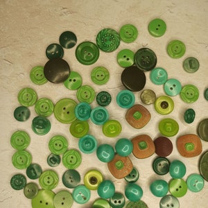 May include: A collection of various green buttons in different shades and sizes. The buttons are scattered across a neutral-toned surface, showcasing a range of textures and designs, from smooth to textured, and round to square.