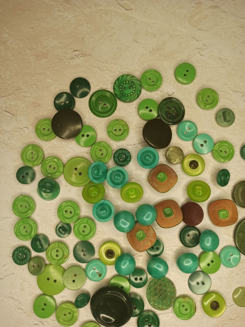 May include: A collection of various green buttons in different shades and sizes. The buttons are scattered across a neutral-toned surface, showcasing a range of textures and designs, from smooth to textured, and round to square.