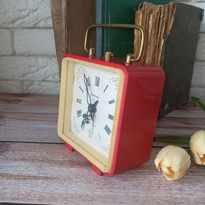 May include: A vintage red and cream-colored alarm clock with a gold handle. The clock face features black Roman numerals and ornate hands. The clock is set against a backdrop of old books and a wooden surface.