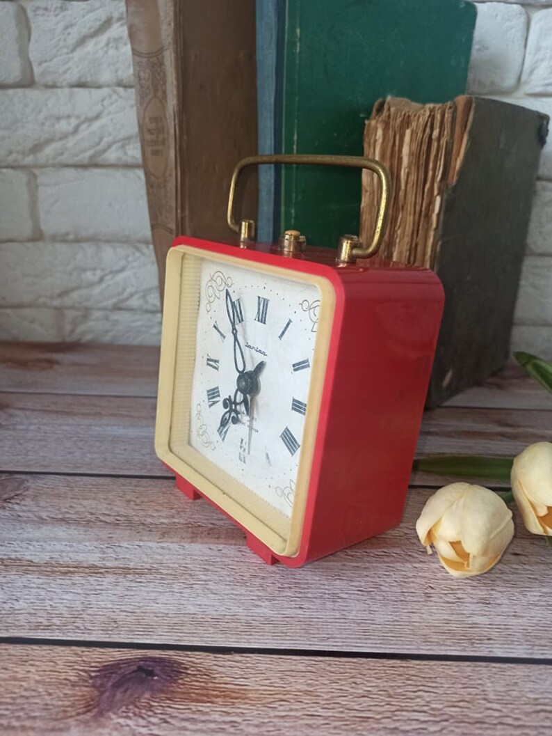 May include: A vintage red and cream-colored alarm clock with a gold handle. The clock face features black Roman numerals and ornate hands. The clock is set against a backdrop of old books and a wooden surface.