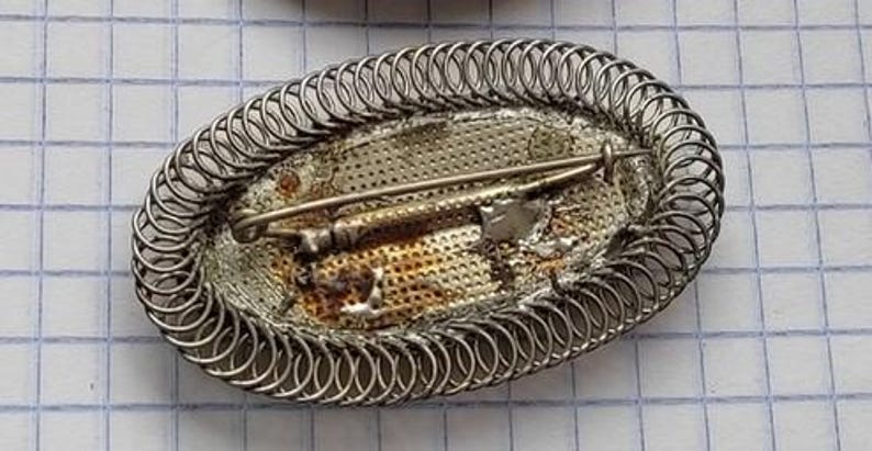 May include: A silver oval brooch with a decorative wire design and a silver pin on the back.
