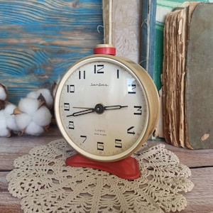 May include: A vintage cream-colored alarm clock with black numbers and hands. The clock face reads "Lantex 4 Jewels Made in USSR". The clock is sitting on a white doily.