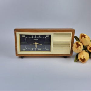 Puede incluir: Un reloj de madera vintage con esfera negra y manecillas doradas. El reloj tiene forma rectangular con un marco color crema y un diseño similar a un altavoz en el lado derecho. El reloj está sobre una superficie blanca.