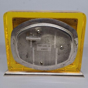May include: A vintage, rectangular clock with a yellow, transparent frame and a silver-colored back. The back of the clock has vertical lines and the text "ГОСТ 3309-58 КЛАСС2". The clock sits on a silver base.