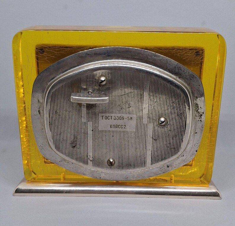 May include: A vintage, rectangular clock with a yellow, transparent frame and a silver-colored back. The back of the clock has vertical lines and the text "ГОСТ 3309-58 КЛАСС2". The clock sits on a silver base.