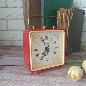 May include: A vintage red and cream-colored alarm clock with a gold handle. The clock face features Roman numerals and decorative flourishes. Several antique books are in the background, suggesting a classic or retro theme.