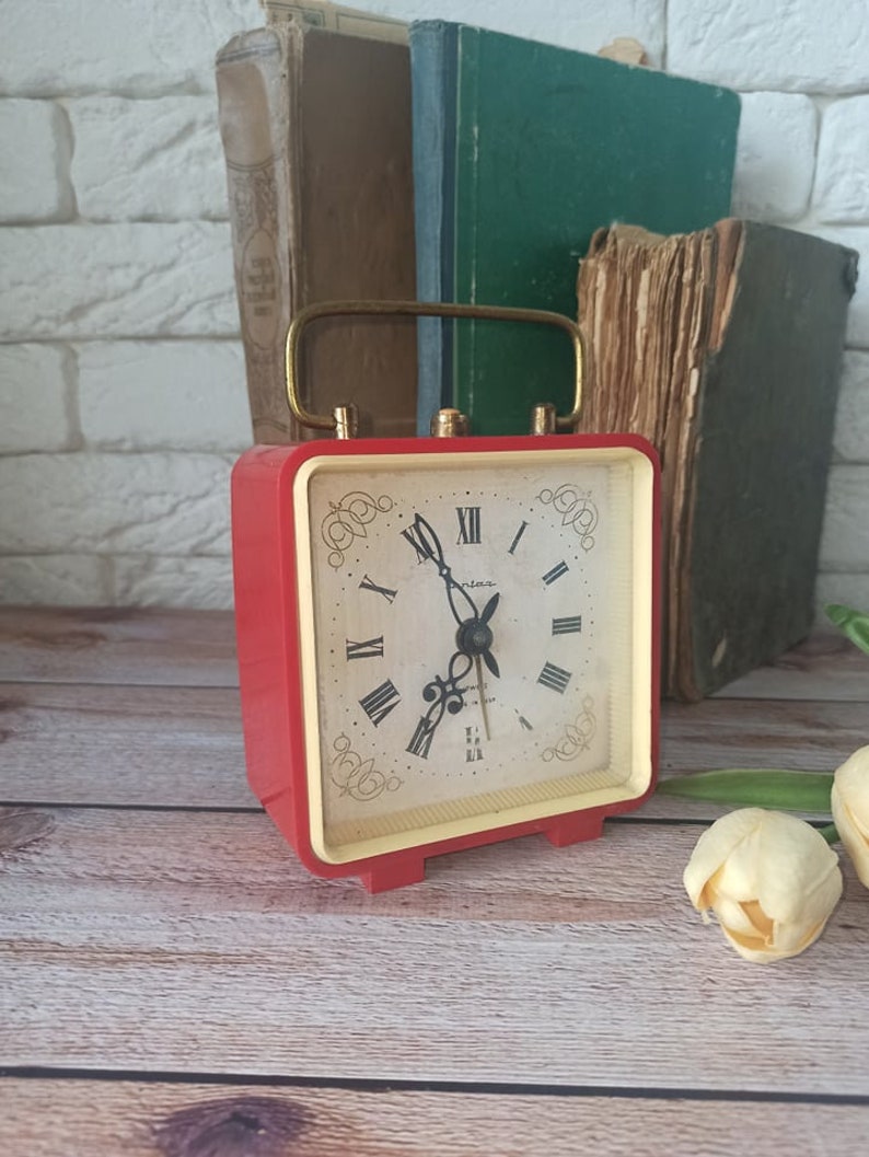 May include: A vintage red and cream-colored alarm clock with a gold handle. The clock face features Roman numerals and decorative flourishes. Several antique books are in the background, suggesting a classic or retro theme.