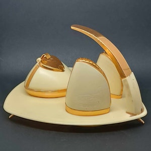 Puede incluir: Un juego de encendedor de cigarrillos vintage blanco y dorado con un soporte curvo y tres compartimentos. El encendedor tiene una tapa dorada y una base blanca. El juego está sobre una superficie blanca.