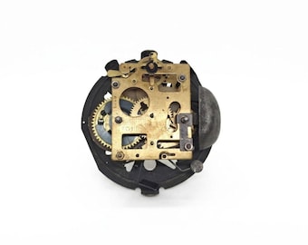 May include: Clock mechanism with brass gears and a black metal frame. The gears are visible, showcasing the intricate internal workings of the clock. The mechanism is set against a white background.