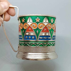 May include: A silver metal cup holder with a green, orange, red, white, and blue geometric design. The cup holder has a handle and a textured base.