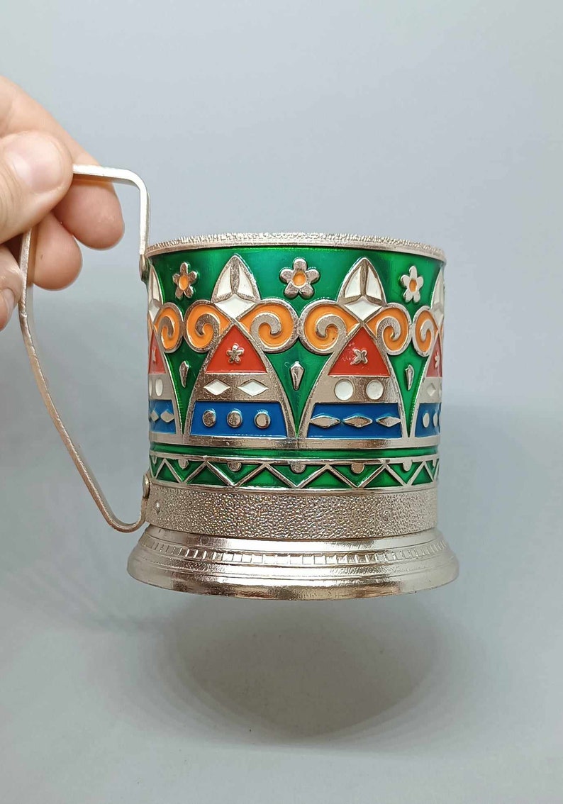 May include: A silver metal cup holder with a green, orange, red, white, and blue geometric design. The cup holder has a handle and a textured base.
