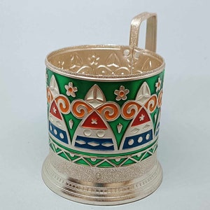 May include: A silver metal cup holder with a green, orange, blue, and white geometric design. The holder has a handle and a textured base.