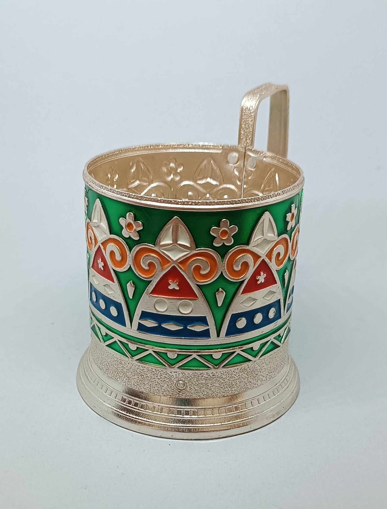 May include: A silver metal cup holder with a green, orange, blue, and white geometric design. The holder has a handle and a textured base.