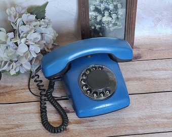 May include: A vintage blue rotary dial telephone with a black dial and handset. The phone is on a wooden surface, with a framed black and white photo and a bouquet of light purple flowers in the background.