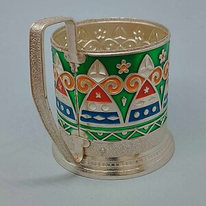 May include: Silver-plated glass holder with a green, orange, blue, and white geometric pattern. The holder has a handle and a decorative base.