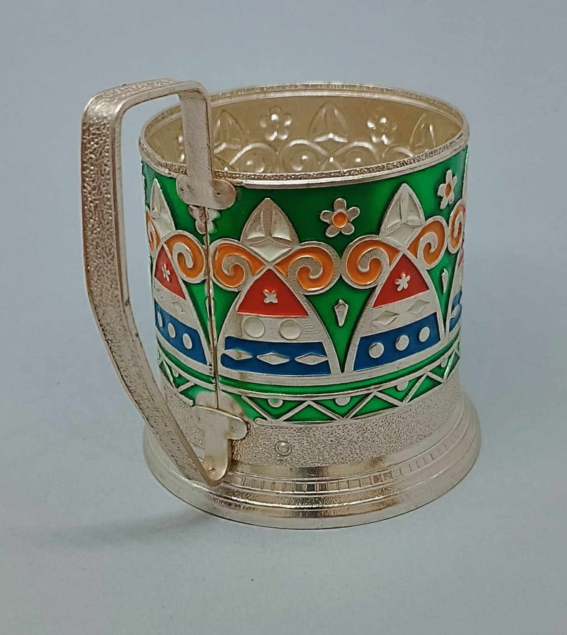 May include: Silver-plated glass holder with a green, orange, blue, and white geometric pattern. The holder has a handle and a decorative base.
