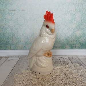 May include: White ceramic cockatoo figurine with a red crest. The bird is perched on a base with a hole in the bottom.