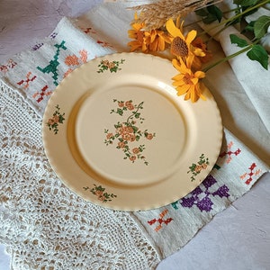 Puede incluir: Un plato crema con borde festoneado, adornado con motivos florales naranjas y verdes. El plato descansa sobre un paño blanco bordado, con flores amarillas y hierba seca. Un objeto decorativo de estilo vintage.
