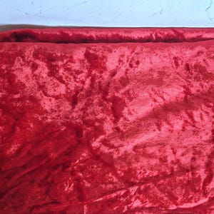 May include: A close-up of a vibrant red velvet fabric. The plush material has a soft, textured appearance with visible folds and shadows, creating a rich, luxurious look. The fabric is likely used for upholstery, clothing, or decorative purposes.