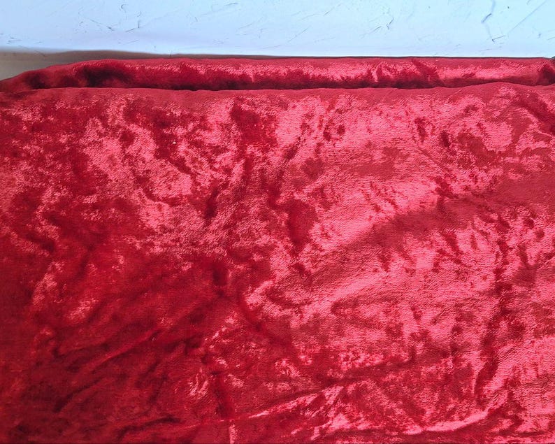 May include: A close-up of a vibrant red velvet fabric. The plush material has a soft, textured appearance with visible folds and shadows, creating a rich, luxurious look. The fabric is likely used for upholstery, clothing, or decorative purposes.