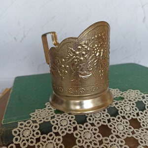May include: A gold-colored metal cup holder with an ornate floral design. The holder has a handle and a scalloped rim.