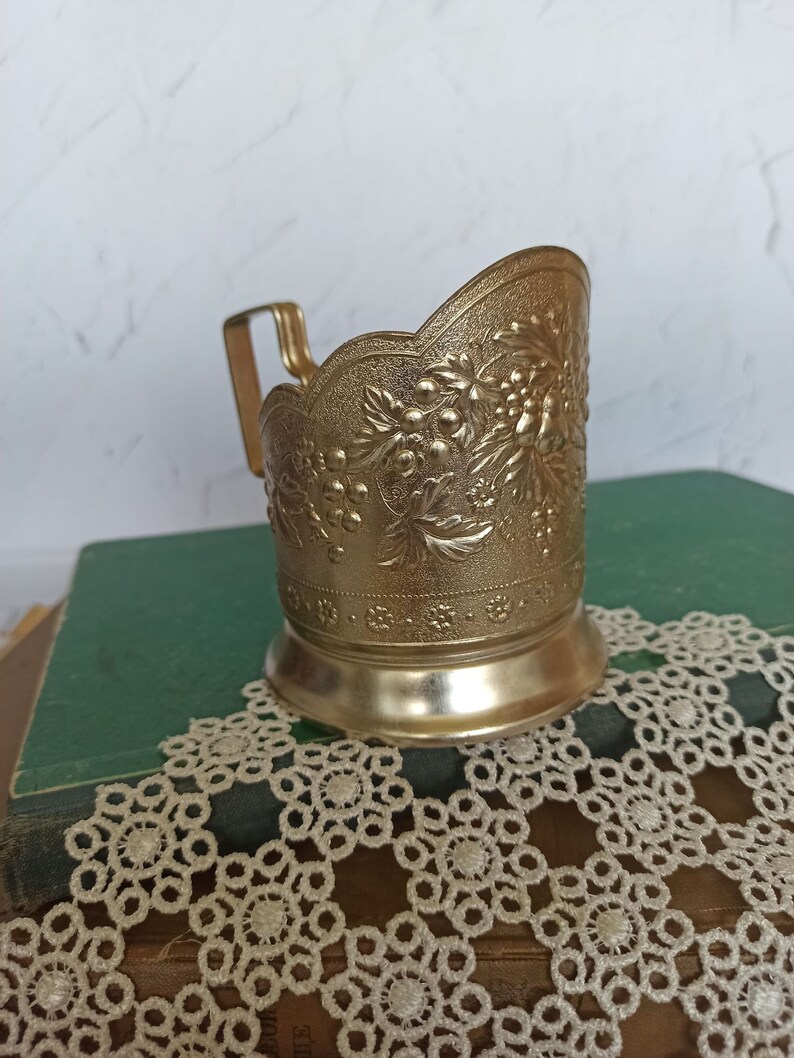 May include: A gold-colored metal cup holder with an ornate floral design. The holder has a handle and a scalloped rim.